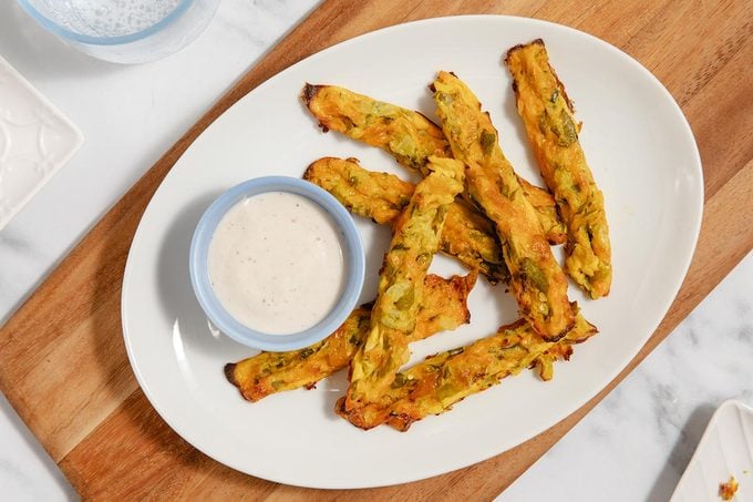 7 cheesy pickle fries on a plate with a ramekin of dipping sauce, on top of a wooden cutting board