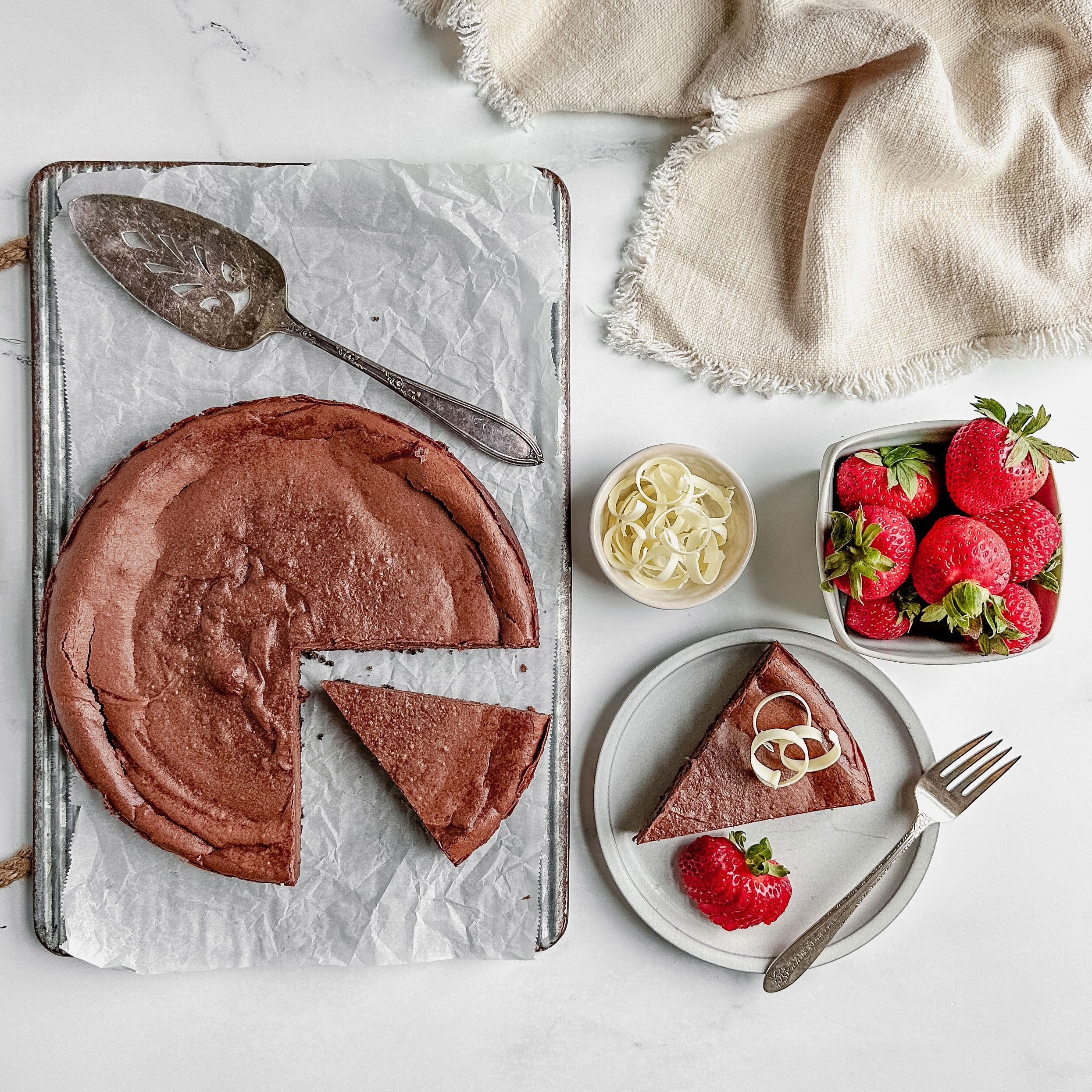 Taste of Home chocolate cheesecake on a white plate with strawberries and white chocolate curls with a linen napkin on a marble surface.