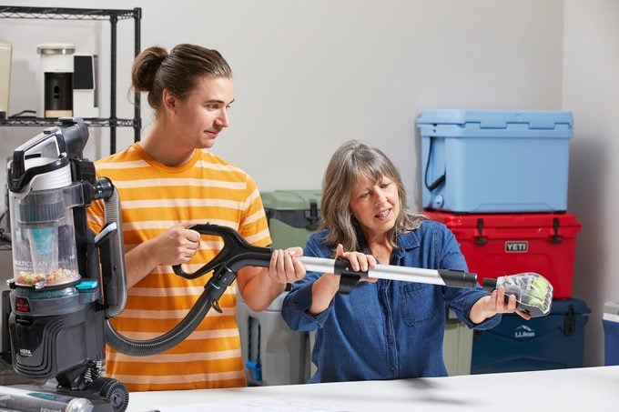 A young man in a striped shirt holds a vacuum cleaner with a long tube, while an older woman in a denim shirt shows him how to use it. They stand by a table in a room with colorful storage boxes in the background.