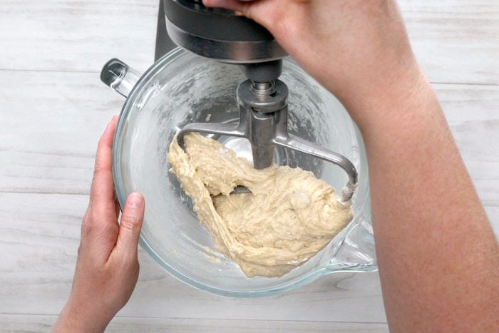 overhead shot of a stand mixer with dough mixing inside; the mixer is in a glass bowl and the dough is a light brown color; a person is holding the bowl with their left hand and the right hand is turning the mixer