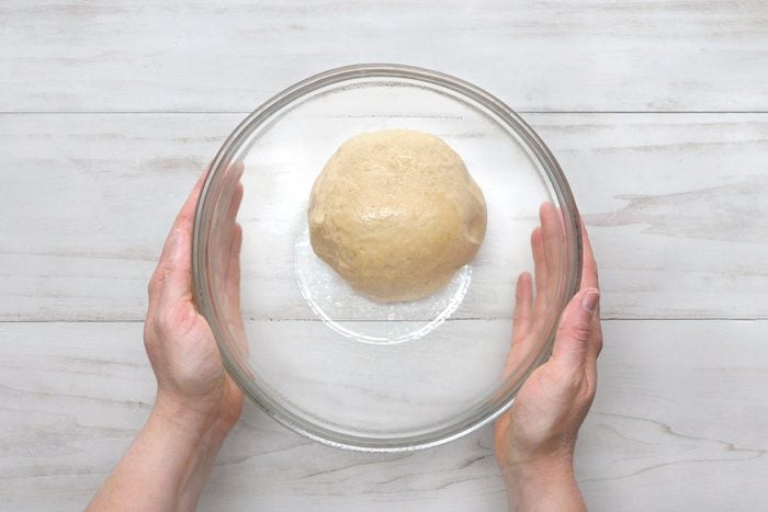 overhead shot of a glass bowl with a ball of dough inside, held by two hands on either side of the bowl; the bowl is on top of a white wooden surface