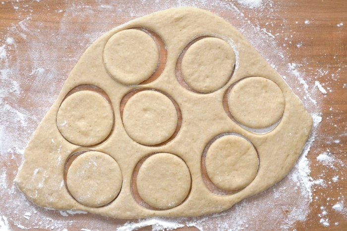 overhead shot of a piece of dough that has been rolled out on a floured wooden surface, it has been cut into seven circles using a cookie cutter; the circles are arranged in a semi-circle shape and there is some flour dusting on the wooden surface and on the dough