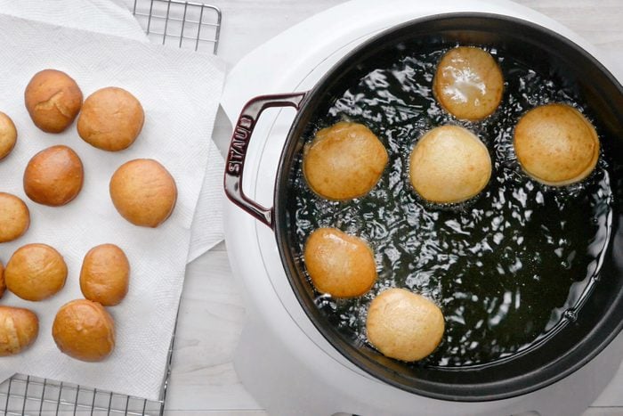 overhead shot of a black cast iron pan with a red handle sits on a white burner, filled with hot oil in which six round, golden-brown donuts are frying; on the left side of the image are twelve donuts of the same type, already fried and laid out on a white paper towel, with a wire cooling rack in the background