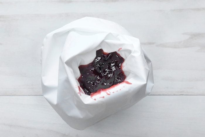 overhead shot of a white piping bag with a red jam-like substance in the bottom; the jam has seeped out a little and is visible on the outside of the bag; the bag is lying on a white wooden surface, likely a table or countertop