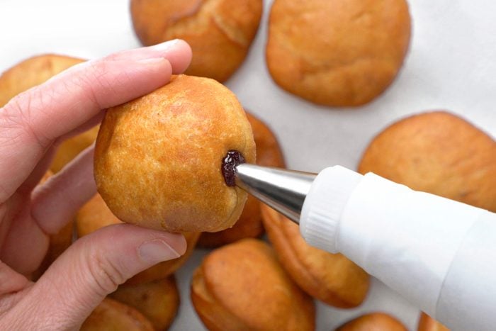 overhead shot of a hand holding a fried dough ball, with a piping bag injecting filling into the hole; a piping bag is a long, narrow, usually white, tube made of cloth or plastic, with a star-shaped or other shaped nozzle at one end