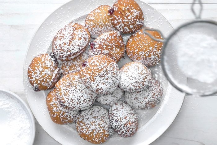 overhead shot of a plate of jelly donuts is shown in this image, covered in powdered sugar; the donuts are piled on top of each other, and some of the jelly is showing through the dough; a sifter filled with powdered sugar is hovering over the donuts, and some of the powder is falling onto the donuts and the plate